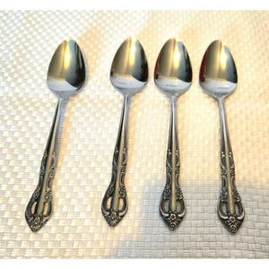 Puede incluir: Cuatro cucharas de metal plateado dispuestas en fila. Cada cuchara presenta un cuenco ovalado pulido y reflectante, y un mango decorativo con intrincados patrones florales y de volutas en relieve en la base y a lo largo del tallo.