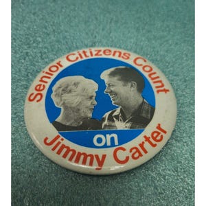 Op de afbeelding: Een ronde campagnebutton met een witte rand en een blauw midden. De button toont afbeeldingen van een oudere vrouw en een man, met de tekst "Senior Citizens Count on Jimmy Carter" in rood en wit.