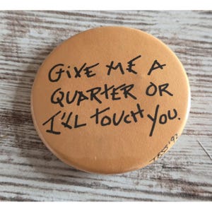 Op de afbeelding: Een ronde, beige-kleurige pinback button met zwarte handgeschreven tekst die luidt: "GIVE ME A QUARTER OR I'LL TOUCH YOU." In de rechterbenedenhoek staat de kleine, handgeschreven handtekening "T&S '92" in zwarte inkt op het oppervlak.