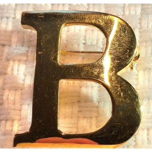 Puede incluir: Un broche de metal en tono dorado con la forma de la letra mayúscula B. La superficie de la letra presenta un acabado pulido y reflectante con reflejos de luz visibles y texturas sutiles en toda la cara frontal del carácter.