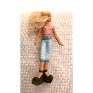 Puede incluir: Una muñeca con cabello rubio largo, que viste una blusa sin mangas de color rosa claro y pantalones cortos de color azul claro. La muñeca tiene extremidades de tono oscuro y lleva zapatos de plataforma negros con suelas gruesas y correas.