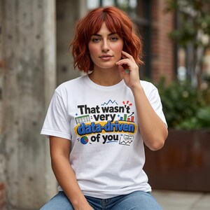 Może przedstawiać: Biały t-shirt z krótkim rękawem z napisem "That wasn't very data-driven of you" czarnymi i niebieskimi literami. Wzór zawiera kolorowe elementy graficzne, takie jak arkusz kalkulacyjny, wykres słupkowy, wykres kołowy i wykres liniowy.