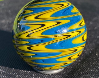 May include: A spherical glass paperweight featuring a swirling pattern of vibrant yellow, deep blue, and thin black lines. The surface is smooth and reflective, showcasing a complex, marbled design that wraps around the entire orb-shaped object.