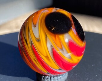 May include: A spherical glass marble featuring a swirling pattern of vibrant orange, bright yellow, and deep red colors. A prominent, solid black circular patch is visible on the upper surface, contrasting with the translucent, multi-colored glass.