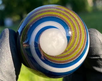 May include: A spherical glass marble featuring a central white core surrounded by concentric swirling patterns of blue, brown, and lime green. The surface of the glass is smooth and reflective, displaying a spiral design that radiates outward from the center.