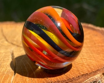 May include: A spherical glass marble featuring swirling bands of orange, red, yellow, and black colors encased within a clear glass orb. The surface of the marble is smooth and reflective, showing internal air bubbles and vibrant color patterns.