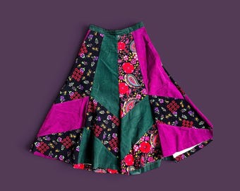 May include: A flared skirt featuring a patchwork design of solid dark green and magenta panels alternating with black fabric panels printed with colorful floral and paisley patterns in shades of red, yellow, and white, finished with a fitted waistband.