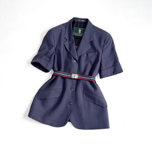 Könnte beinhalten: Ein marineblauer Blazer mit kurzen Ärmeln und einem mehrfarbigen Streifengürtel. Der Blazer hat ein klassisches Design mit einem Reverskragen, zwei Vordertaschen und Knopfverschlüssen. Der Gürtel hat rote, blaue und schwarze Streifen. Der Blazer ist Größe Medium.