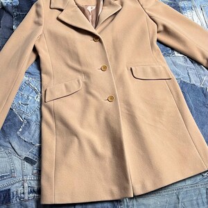 May include: A tan coat with a classic design. The coat has a notched collar, three buttons, and two front pockets. The coat is made of a soft material and has the brand name "DAMO" on the label.