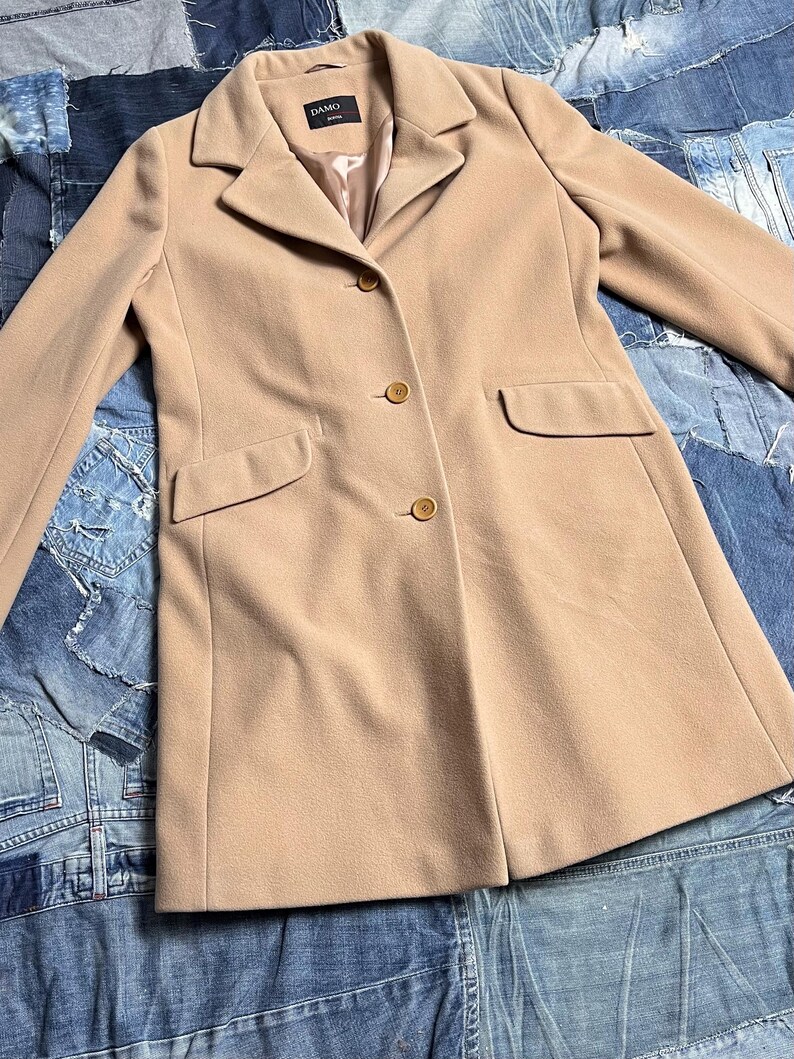May include: A tan coat with a classic design. The coat has a notched collar, three buttons, and two front pockets. The coat is made of a soft material and has the brand name "DAMO" on the label.