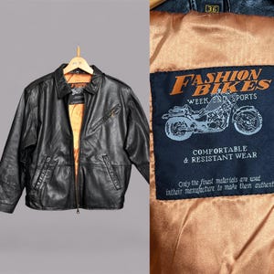 May include: Black leather jacket with a collared neckline and a front zipper closure. The jacket has two front pockets and a small zippered pocket on the chest. The interior lining is a golden-brown color. The label reads "Fashion Bikes Week End Sports."