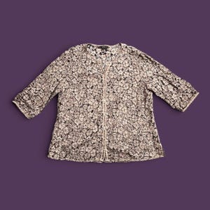 May include: A beige and black floral lace cardigan featuring three-quarter length sleeves, a round neckline, and a vertical front button closure. The garment is constructed from a semi-sheer, intricate lace fabric with a repeating floral pattern.