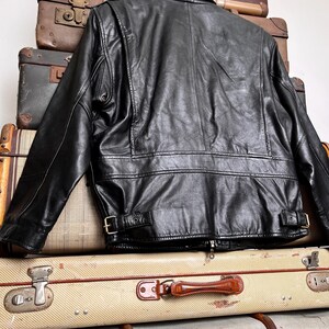 May include: Black leather jacket with a classic design, featuring a collar, zipper, and buckle details. The jacket is displayed on top of a stack of vintage suitcases, showcasing a retro aesthetic.