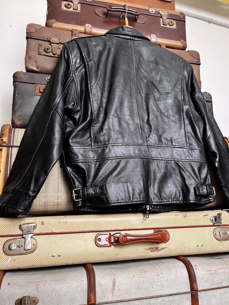 May include: Black leather jacket with a classic design, featuring a collar, zipper, and buckle details. The jacket is displayed on top of a stack of vintage suitcases, showcasing a retro aesthetic.