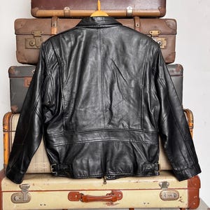 May include: Black leather jacket with a classic design, featuring a collar, zipper, and belt details. The jacket is displayed on a wooden hanger, set against a backdrop of vintage suitcases. A timeless piece for any wardrobe.