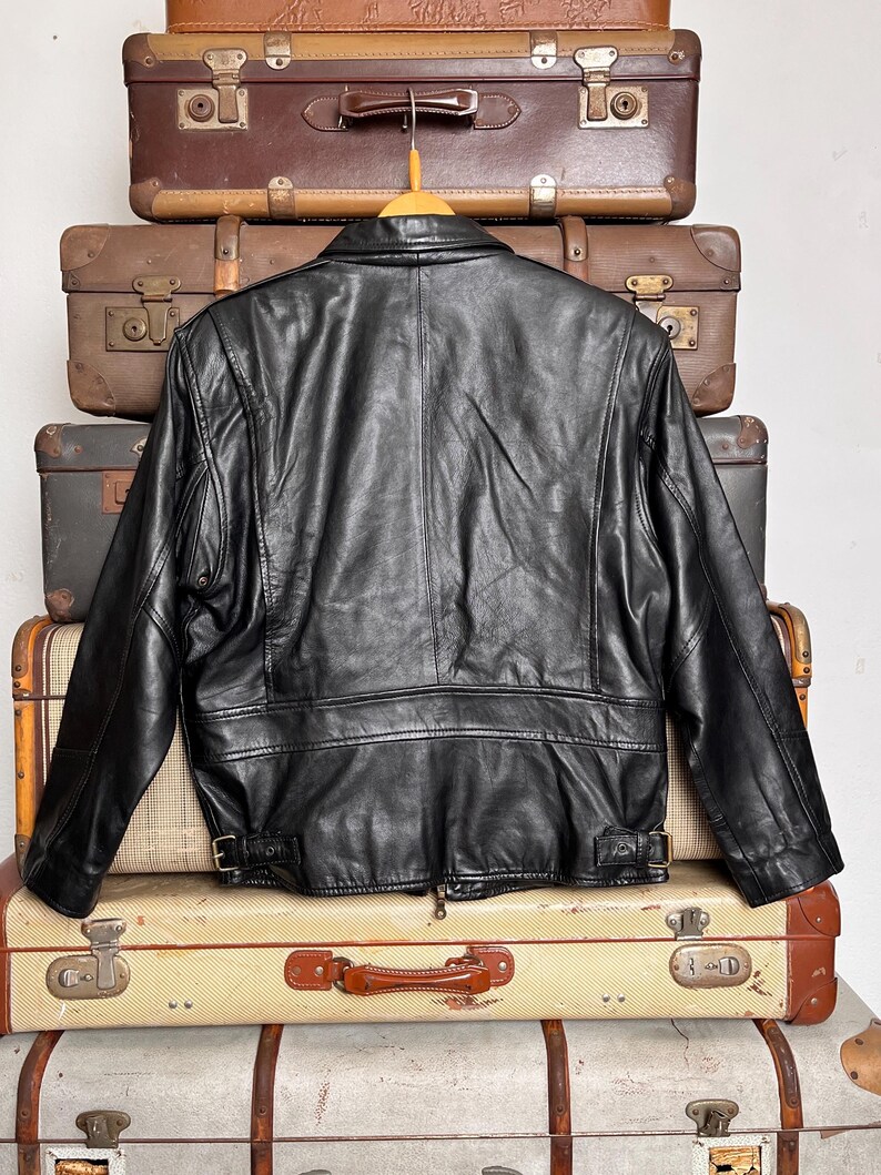 May include: Black leather jacket with a classic design, featuring a collar, zipper, and belt details. The jacket is displayed on a wooden hanger, set against a backdrop of vintage suitcases. A timeless piece for any wardrobe.