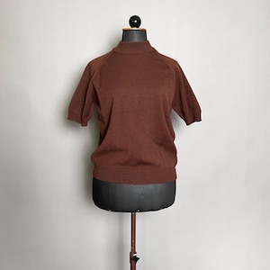 以下が含まれることがあります： ブラウンの半袖ニットトップス、ハイネック。 黒いマネキンに展示されています。 リブ編みの裾と袖口があります。 マネキンは茶色の金属スタンドで支えられています。