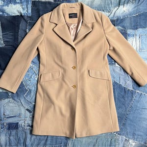 May include: A beige coat with a tailored design. The coat has a notched lapel, two front pockets, and three buttons. The coat is made of a soft fabric and has the brand name "DAMO" on a label.
