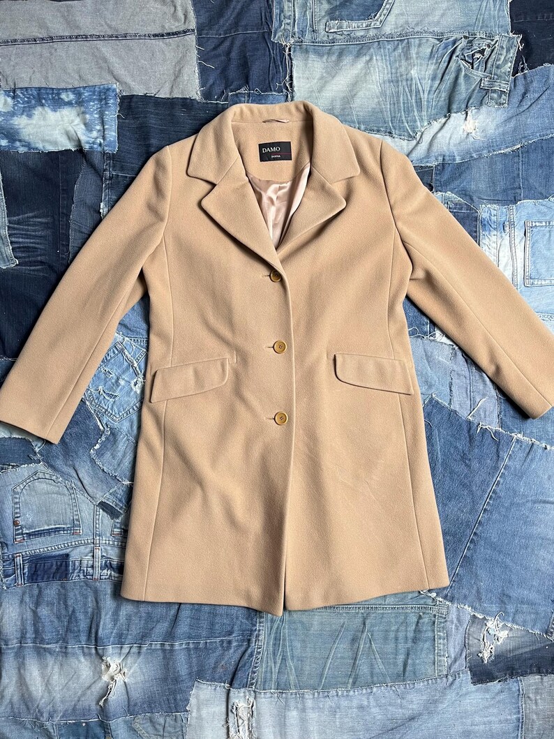 May include: A beige coat with a tailored design. The coat has a notched lapel, two front pockets, and three buttons. The coat is made of a soft fabric and has the brand name "DAMO" on a label.