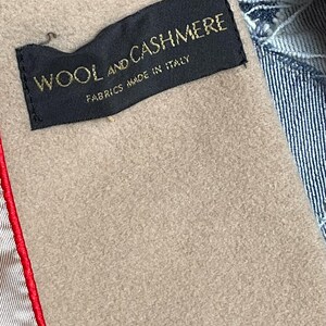 May include: A close-up shot of a black label with gold text reading "WOOL AND CASHMERE FABRICS MADE IN ITALY". The label is stitched into a tan fabric, with a red seam on the left and denim on the right.