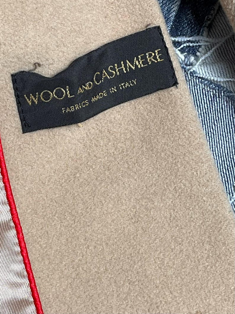 May include: A close-up shot of a black label with gold text reading "WOOL AND CASHMERE FABRICS MADE IN ITALY". The label is stitched into a tan fabric, with a red seam on the left and denim on the right.