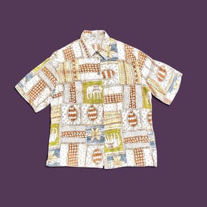May include: A short-sleeve button-down shirt featuring a patchwork pattern of squares. The design includes illustrations of elephants, flowers, and geometric shapes in shades of brown, olive green, blue, and white on a light background.