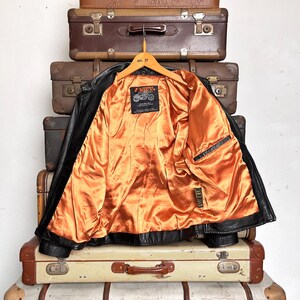 May include: Black leather jacket with an orange satin lining, hanging on a wooden hanger. The jacket is displayed against a backdrop of vintage brown suitcases. The jacket has a classic motorcycle style with a notched collar and a zipper closure.