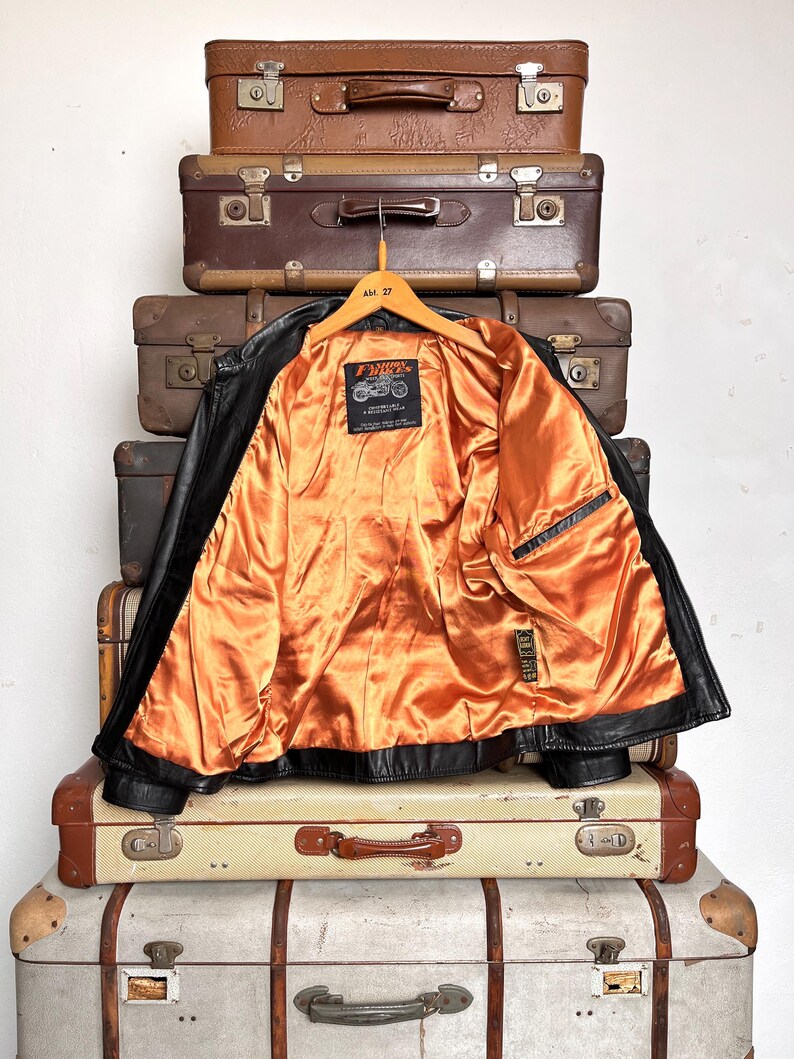 May include: Black leather jacket with an orange satin lining, hanging on a wooden hanger. The jacket is displayed against a backdrop of vintage brown suitcases. The jacket has a classic motorcycle style with a notched collar and a zipper closure.