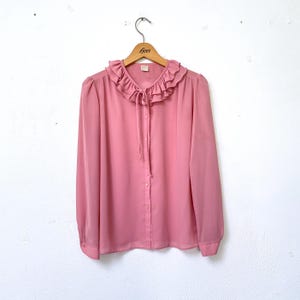 May include: A pink long-sleeved blouse with a ruffled collar and a tie at the neck. The blouse is buttoned down the front.