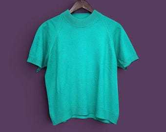May include: A short-sleeved knit sweater in a vibrant turquoise green color. The garment features a high mock neck collar, ribbed detailing along the neckline, sleeve cuffs, and bottom hem, and raglan-style seams extending from the neck to the armpits.