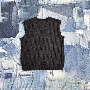 Op de afbeelding: Een zwarte mouwloze trui met een V-hals. De trui heeft een textuur, open gebreid patroon en is geplaatst op een patchwork denim achtergrond.