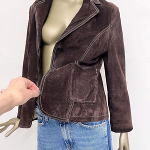 Puede incluir: Una chaqueta de ante marr&oacute;n con costuras blancas. La chaqueta tiene un cierre de bot&oacute;n &uacute;nico y un bolsillo de parche en el lado izquierdo.
