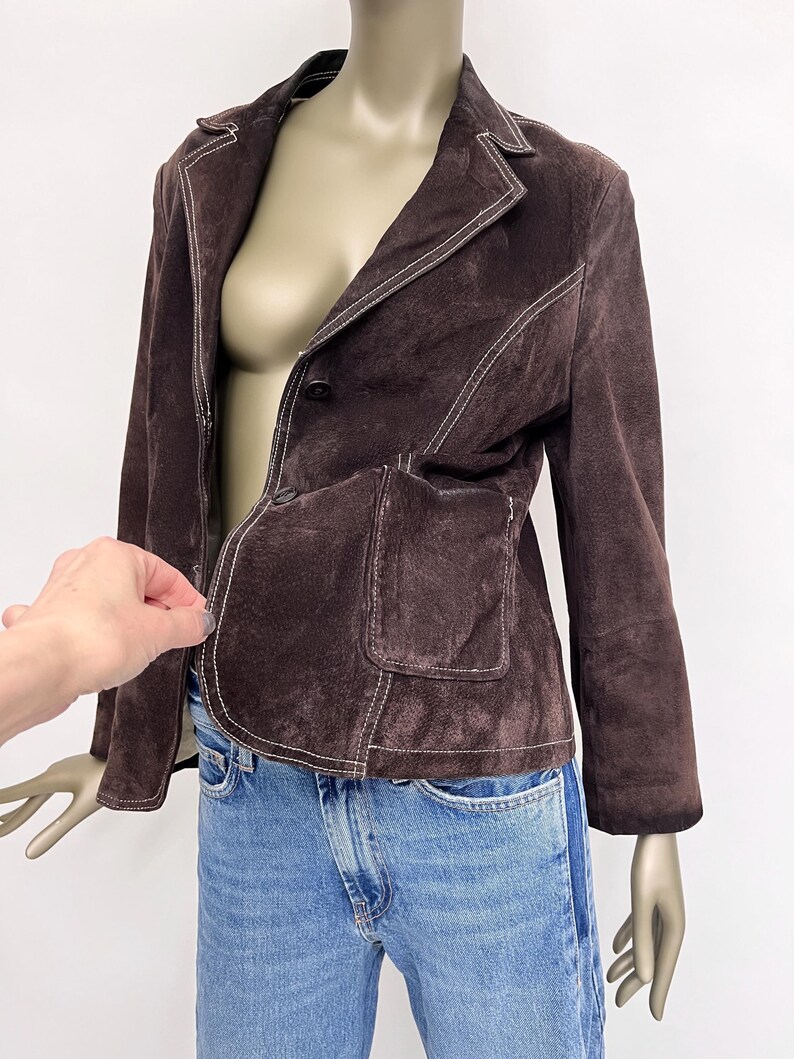 Puede incluir: Una chaqueta de ante marr&oacute;n con costuras blancas. La chaqueta tiene un cierre de bot&oacute;n &uacute;nico y un bolsillo de parche en el lado izquierdo.