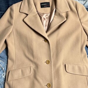May include: A beige coat with a traditional design. The coat has a notched collar, three buttons, and two front pockets. The label reads "DAMO DONNA".