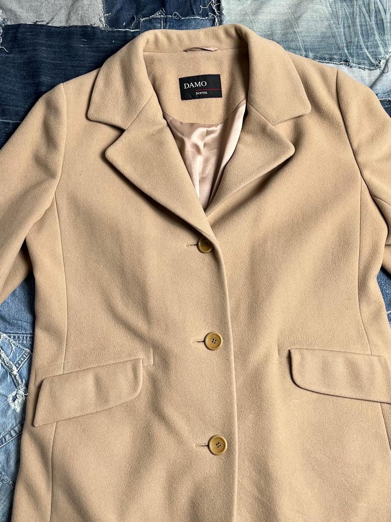 May include: A beige coat with a traditional design. The coat has a notched collar, three buttons, and two front pockets. The label reads "DAMO DONNA".
