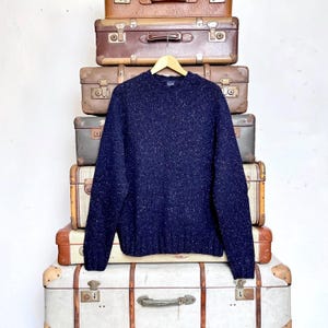 Könnte beinhalten: Ein marineblauer Pullover mit gesprenkeltem Muster, präsentiert auf einem Holzbügel. Der Pullover hat einen Rundhalsausschnitt und lange Ärmel. Im Hintergrund ein Stapel Vintage-Koffer in verschiedenen Größen und Farben.