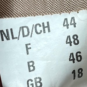 May include: A white clothing label with black text showing size designations for NL/D/CH, F, B, and GB, with corresponding numbers 44, 48, 46, and 18. The label is set against a textured brown fabric.