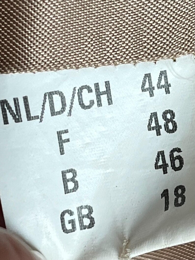 May include: A white clothing label with black text showing size designations for NL/D/CH, F, B, and GB, with corresponding numbers 44, 48, 46, and 18. The label is set against a textured brown fabric.