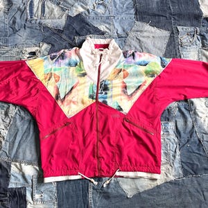 May include: A vintage magenta windbreaker jacket with a full zipper and a white collar. The jacket features a colorful geometric pattern across the shoulders and chest. It has two zippered pockets and a drawstring at the waist.