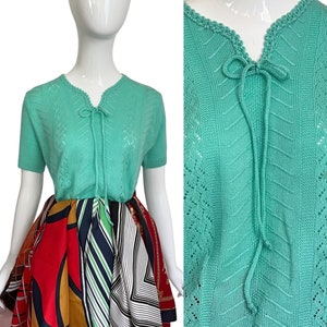 May include: A mint green knitted sweater with a V-neck and a tie at the neckline. The sweater has a delicate openwork pattern.