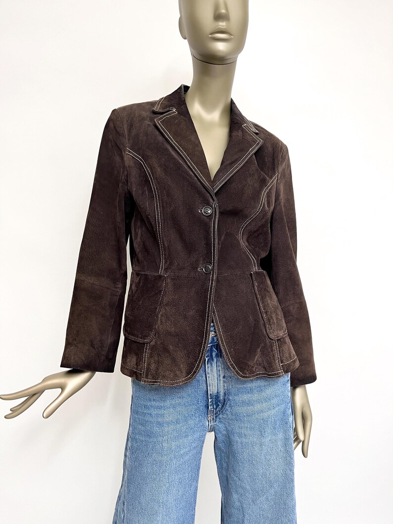 Puede incluir: Una chaqueta de ante marr&oacute;n con costuras blancas. La chaqueta tiene un cuello de muesca, cierre de bot&oacute;n y dos bolsillos de solapa.