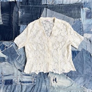 Può includere: Top in pizzo avorio con maniche corte e scollo a V. Il top è abbottonato sul davanti e ha un orlo smerlato. Il top è esposto su uno sfondo di denim patchwork.