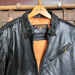 May include: Black leather jacket with a zip-up front, a collared neckline, and a small zippered chest pocket. The jacket has an orange lining and a label that reads "Fashion Bikes Week & Sports". The jacket is hanging on a wooden hanger.