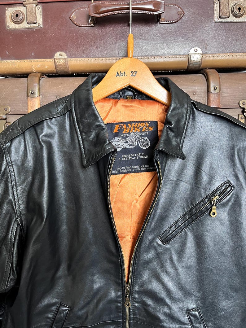 May include: Black leather jacket with a zip-up front, a collared neckline, and a small zippered chest pocket. The jacket has an orange lining and a label that reads "Fashion Bikes Week & Sports". The jacket is hanging on a wooden hanger.