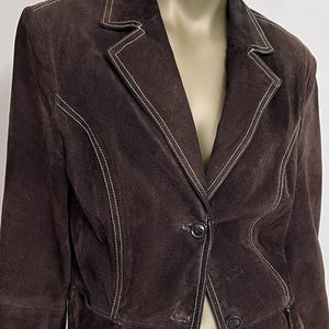 Puede incluir: Una chaqueta de ante marr&oacute;n con costuras blancas. La chaqueta tiene un cierre de bot&oacute;n &uacute;nico y dos bolsillos.