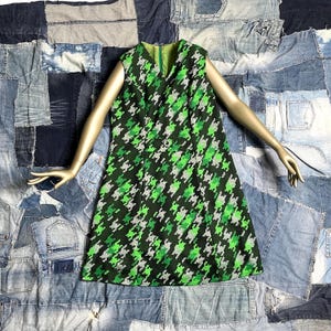Könnte beinhalten: Ein ärmelloses Kleid im Vintage-Stil mit einem grün-schwarz-weißen Hahnentrittmuster. Das Kleid hat einen V-Ausschnitt und ein Knopfdetail. Es wird auf einem Schaufensterpuppenarm vor einem Patchwork-Denim-Hintergrund präsentiert.