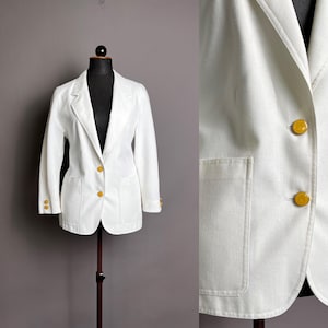 May include: A white blazer with gold buttons and two front pockets. The blazer is displayed on a black mannequin. The blazer has long sleeves and a notched lapel. The buttons are round and have a gold color.