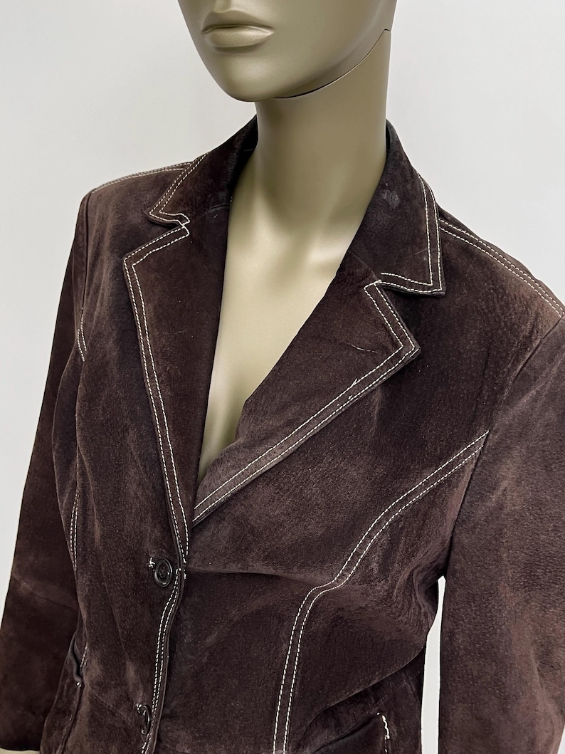 Puede incluir: Una chaqueta de ante marr&oacute;n con costuras blancas. La chaqueta tiene un cierre de bot&oacute;n &uacute;nico y un cuello.