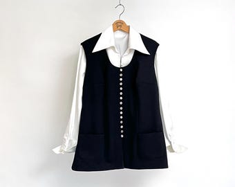 May include: A black sleeveless dress with a button-up front and two pockets. The dress is worn over a white collared shirt.