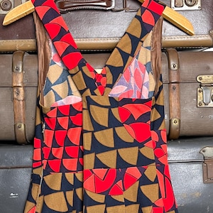 May include: A sleeveless dress featuring a geometric pattern of semicircles in red, tan, and black. The garment has a V-neckline, wide shoulder straps, and a side zipper closure. The interior is lined with a solid brown fabric, and the dress is displayed on a wooden hanger.